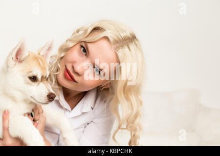 Donna bionda con cucciolo di cane husky su un divano bianco. ragazza che gioca con un cane Foto Stock