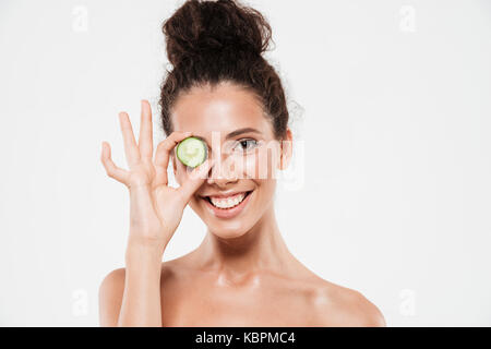 Ritratto di bellezza di un sorridente giovane donna con morbida pelle sana azienda fetta di cetriolo presso il suo occhio isolate su sfondo bianco Foto Stock