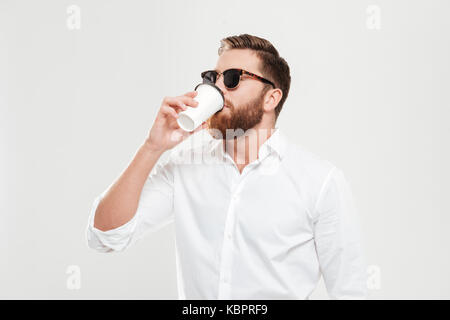 Immagine del bel giovane uomo barbuto in piedi su parete bianca isolata di sfondo di bere il caffè. Guardando a parte. Foto Stock