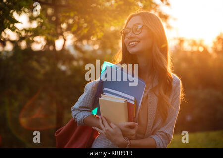 Sorridente brunette donna in occhiali da vista che abbraccia i libri e guardando lontano in posizione di parcheggio Foto Stock