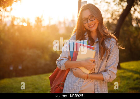Sorridente brunette donna in occhiali da vista che abbraccia i libri e guardando la telecamera in posizione di parcheggio Foto Stock
