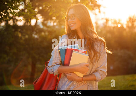 Happy brunette donna in occhiali da vista che abbraccia i libri e guardando lontano in posizione di parcheggio Foto Stock