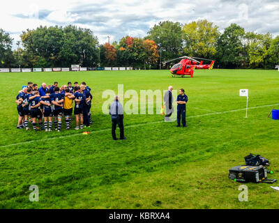 Woodford Green, Londra, Regno Unito. Il 30 settembre 2017. I giocatori conferiscono come London Air Ambulance elicotteri G-LNDN atterra a Woodford Rugby Football Club Highams massa nel Woodford Green in risposta a una relazione di uno spettatore la sofferenza di un attacco di cuore. Il Woodford v Chelmsford match è stato sospeso per 40 minuti mentre i paramedici hanno partecipato alla vittima che era in grado di uscire dalla strada. Woodford è andato a vincere la partita. La Air Ambulance è supportato da Londra massoni. Credito: Mark Dunn/ Alamy Live News Foto Stock