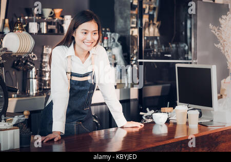 Femmina asiatica barista indossare jean grembiule mettere la sua mano sul banco bar e sorridente al cliente,servizio di caffetteria concetto,proprietario business start up. Foto Stock