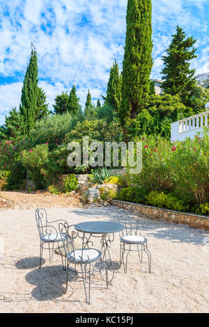 Tre sedie in metallo con tavolo sulla ghiaia patio nel giardino mediterraneo vicino alla città di Primosten, Dalmazia, Croazia Foto Stock