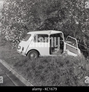 Anni sessanta, storico, un fracassato Mini auto a seguito di un incidente stradale si trova in un fosso accanto a una strada di campagna, Aylesbury, Inghilterra, Regno Unito. Foto Stock