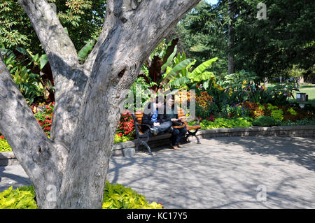 Due persone sedute su una panchina in un parco della città con giardini in background Foto Stock