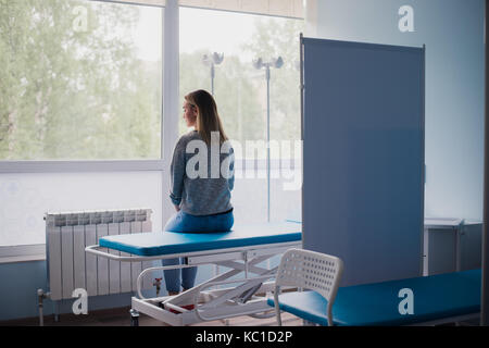 Giovane donna incinta seduta sul letto in confortevoli ward, in attesa del medico premurosamente Foto Stock