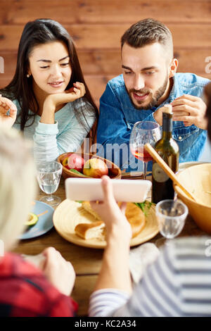 Ritratto di giovane donna asiatica e l uomo moderno guardando lo schermo dello smartphone per discutere le foto mentre a cena con gli amici seduti al grande tavolo con Foto Stock