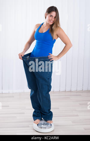 Slim donna in jeans oversize permanente sulla macchina di pesatura a casa Foto Stock