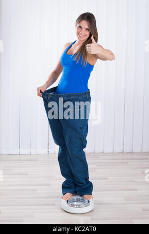 Slim donna in jeans oversize permanente sulla macchina di pesatura a casa Foto Stock