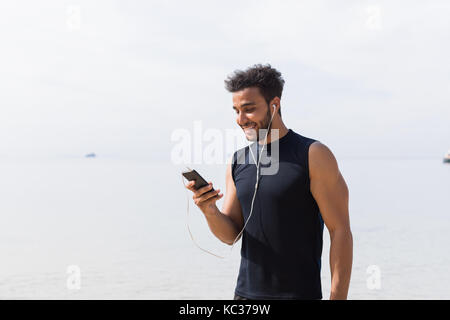 Maschio ispanica runner utilizzando una cella smart phone sulla spiaggia mentre il jogging sul mare uomo giovane sorridenti sport latina Foto Stock