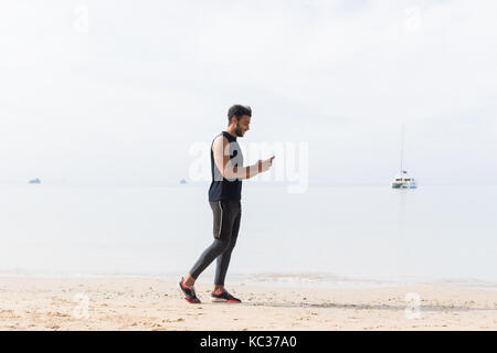 A piena lunghezza shot dei maschi di runner utilizzando una cella smart phone sulla spiaggia mentre il jogging sul mare uomo giovani latino-sport Foto Stock