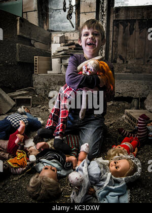 Foto di un inquietante giovane ragazzo in possesso di un vecchio clown bambola in una vecchia stalla coperta in spiderwebs e polvere. Foto Stock