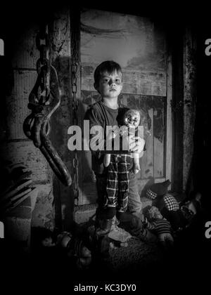 Foto di un inquietante giovane ragazzo in possesso di un vecchio clown bambola in una vecchia stalla coperta in spiderwebs e polvere. Foto Stock