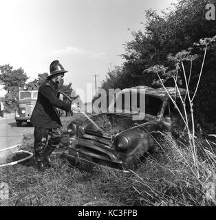 Anni sessanta, storico allarmi antincendio uniforme che indossano il casco e la spruzzatura di acqua in un bruciate le auto di piccole dimensioni su un orlo a lato di una strada di campagna, Inghilterra, Regno Unito. Foto Stock