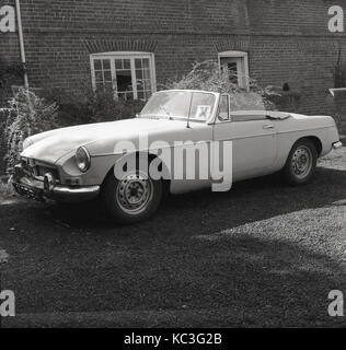 1963, storico, un MGB auto sportiva con i mozzi ruota parcheggiato al di fuori di una casa con la lettera X bloccato sulla finestra, Inghilterra, Regno Unito. Foto Stock