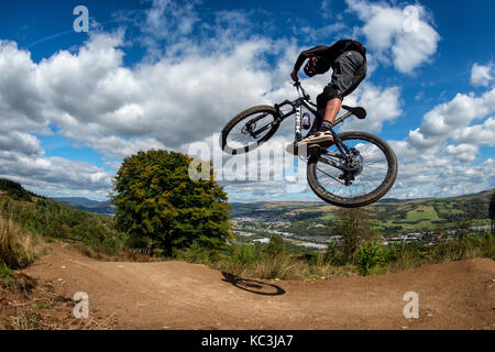 Un mountain biker salta mentre cavalcate un sentiero a Bikepark Galles che si affaccia sulla città di Merthyr Tydfil e il Brecon Beacons all'orizzonte. Foto Stock