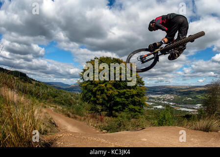 Un mountain biker salta mentre cavalcate un sentiero a Bikepark Galles che si affaccia sulla città di Merthyr Tydfil e il Brecon Beacons all'orizzonte. Foto Stock