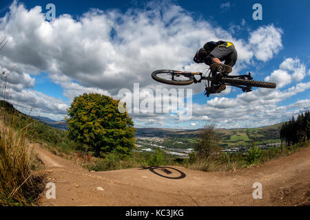 Un mountain biker salta mentre cavalcate un sentiero a Bikepark Galles che si affaccia sulla città di Merthyr Tydfil e il Brecon Beacons all'orizzonte. Foto Stock