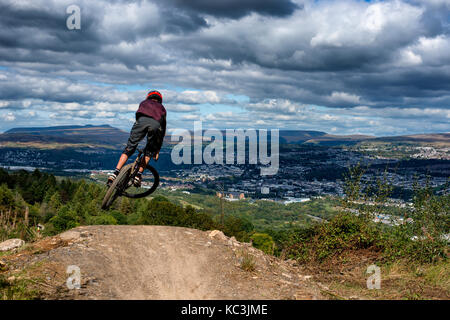 Un mountain biker salta mentre cavalcate un sentiero a Bikepark Galles che si affaccia sulla città di Merthyr Tydfil e il Brecon Beacons all'orizzonte. Foto Stock