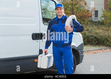 Ritratto di fiducia elettricista con la bobina del cavo e la casella degli strumenti in piedi al di fuori del carrello Foto Stock
