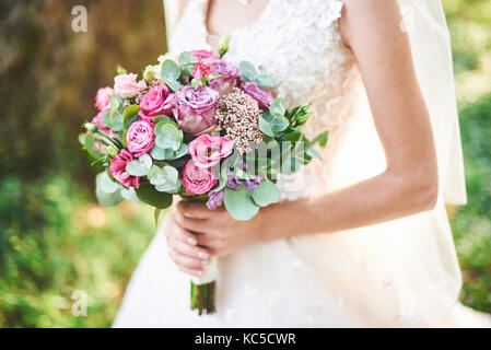 Sposa in un abito bianco tenendo un mazzo di fiori viola e verde su uno sfondo di erba verde Foto Stock