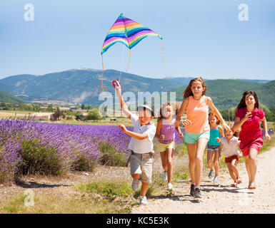 Ragazzo eccitato battenti rainbow kite mentre i suoi amici in esecuzione dopo di lui attraverso il prato di lavanda Foto Stock