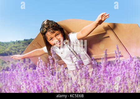 Carino boy vestito come un pilota con ali di cartone volando a bassa quota sopra il prato di lavanda Foto Stock