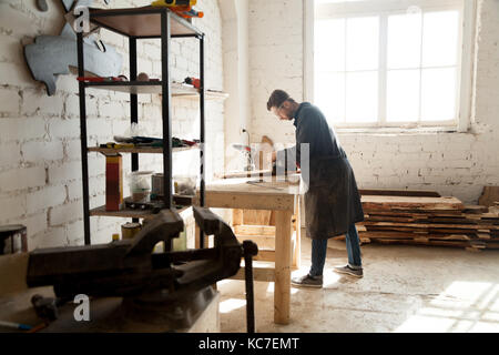 Vista laterale del giovane professionista artigiana della lavorazione del legno nel negozio di piallatura garage. artigiano lavorando su mobili su misura, utilizzando sander. falegnameria Foto Stock