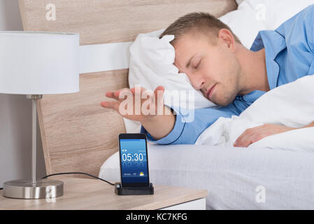 Uomo disteso sul letto del posticipo allarme sul display del cellulare Foto Stock