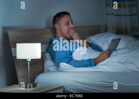 Assonnato uomo sbadigliare sul letto guardando al laptop Foto Stock