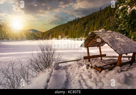 Pergolato in legno in inverno nevoso foreste di abete rosso. bellissimo paesaggio montuoso vicino coperta di neve lago ghiacciato al tramonto Foto Stock