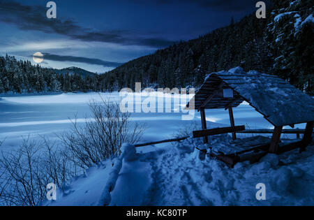 Pergolato in legno in inverno nevoso foreste di abete rosso. bellissimo paesaggio montuoso vicino coperta di neve lago ghiacciato di notte nella luce della luna piena Foto Stock