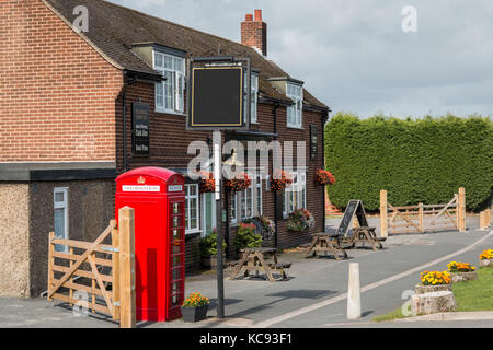 Public House con un segno bianco per inserire testo, cancelli in legno e stile vecchio telefono rosso scatola, fiori in cestelli appesi sulla parete siepe verde in Foto Stock