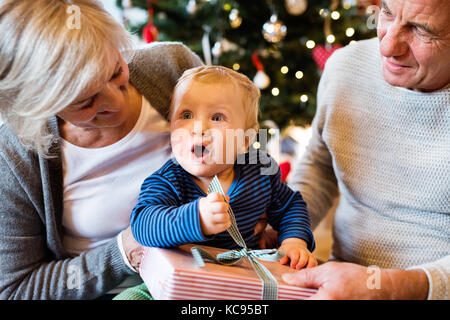 Nonni con il loro piccolo nipote ad albero di Natale a casa presenta il disimballaggio. Foto Stock