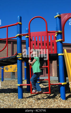 Bambina si arrampica la scaletta del parco giochi alla sua scuola. apparecchiature è in metallo verniciato di rosso e di blu. Foto Stock