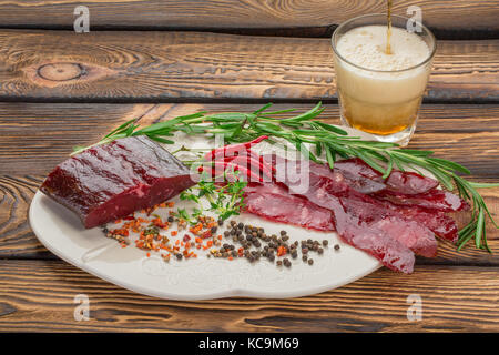 La carne secca con erbe fresche, peperoncino, condimenti su una piastra. La birra viene versata in un bicchiere su sfondo di legno. La birra con schiuma Foto Stock