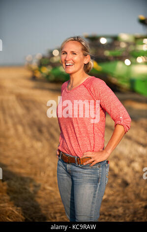 Un giovane agricoltore femmina sorge in un campo vicino a John Deere per le mietitrebbie precedenti per la mietitura del frumento sulla famiglia agriturismo vicino a Breckenridge, North Dakota Foto Stock