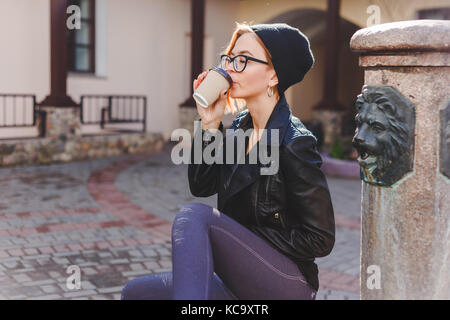 Giovani elegantemente vestito donna in nero berretto lavorato a maglia e bicchieri godendo il caffè dalla carta take away cup. rompere il concetto di tempo Foto Stock