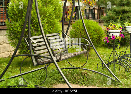 Metallo panchina da giardino-swing nel cortile antistante la recinzione tra cespugli ornamentali e fiori Foto Stock