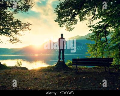 Ragazzo in giacca rossa e pantaloni neri stand sul ceppo di albero. vuota panca di legno al lago di montagna. banca sotto faggi tree, montagne all orizzonte e in wa Foto Stock