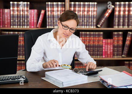 Revisore femmina analizzando le bollette con lente di ingrandimento e calcolatrice sulla scrivania in legno Foto Stock