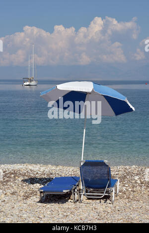 Due sdraio o lettini per prendere il sole su di una tegola o spiaggia di ciottoli sull'isola greca di Corfu con una pantina parasole o ombrello sopra per proteggere dal calore. Foto Stock