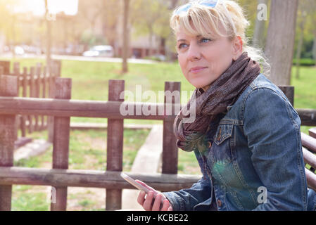 Attraente donna bionda con un mobile seduta su una panchina in un parco riflessivo Foto Stock