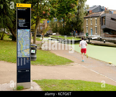 Un pareggiatore corre sul alzaia passando un cartello leggibile della London Column Street al Mile End Lock sul Regents Canal, Londra Foto Stock