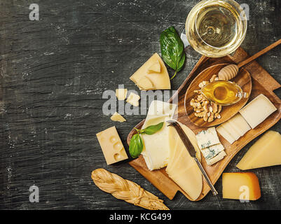 Vari tipi di formaggio sul vassoio in legno con vino bianco su sfondo nero, vista dall'alto Foto Stock
