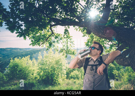 Uomo che indossa gli occhiali da sole e zainetto in appoggio sotto l'albero e acqua potabile Foto Stock