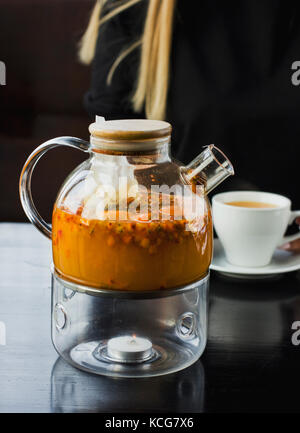 Bacche di olivello spinoso tè in una teiera in vetro. Sullo sfondo di una giovane ragazza bionda bere il tè in cafe Foto Stock