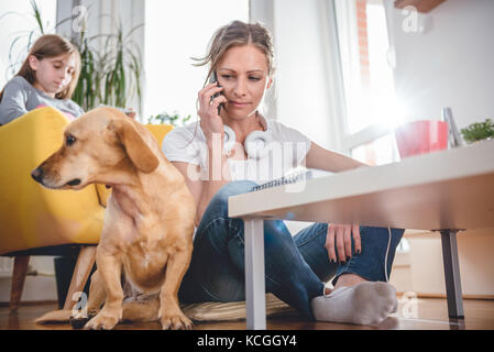 Donna seduta sul pavimento da small yellow dog e parlando al telefono intelligente a casa mentre la figlia seduto in poltrona di colore giallo in background Foto Stock